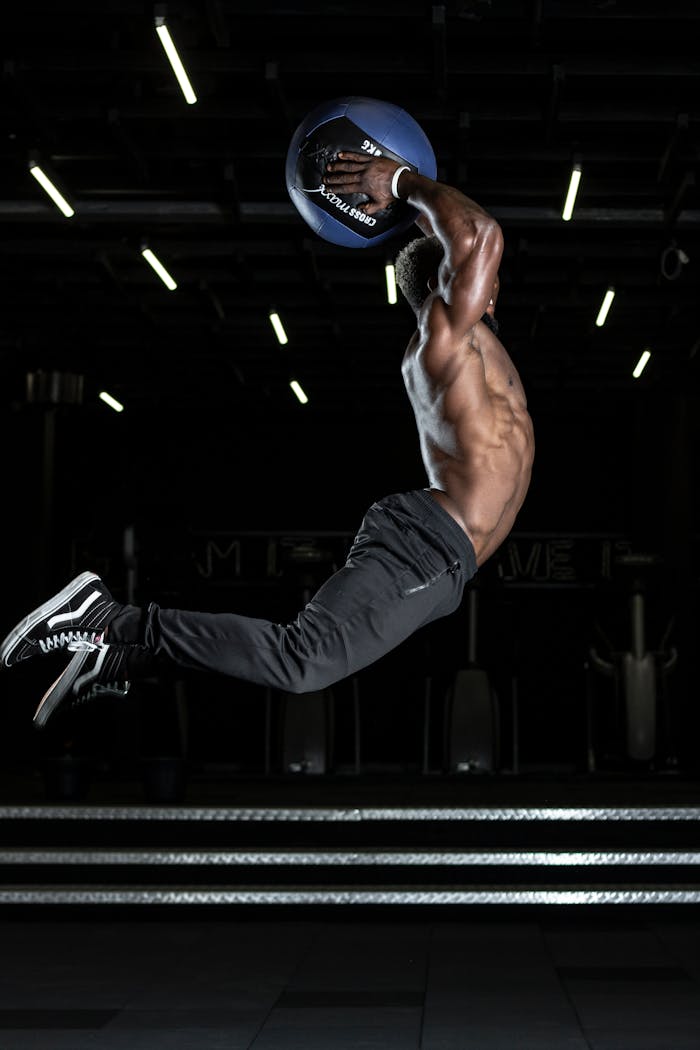 who-we-are Muscular athlete performing a high jump with a medicine ball in a gym, showcasing strength and fitness.